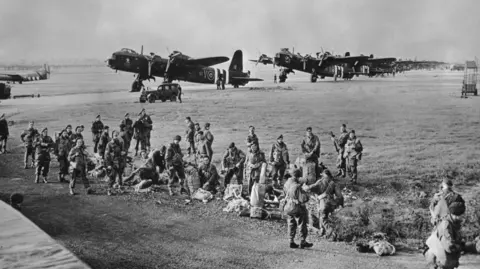 Getty Images An old black and white photo from September 1944 during World War Two. There is a group of soldiers on a field preparing their parachutes and equipment before taking flight for Operation Market Garden in the Netherlands. There are several planes in the distance.