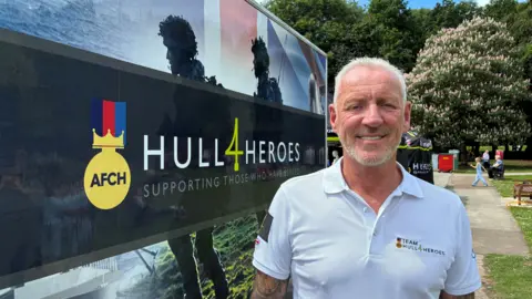 A head and shoulders shot of Paul Matson. He has grey hair and grey facial hair. He is wearing a white polo T-shirt and is stood next to a black sign reading "HULL 4 HEROES" in white capital letters. He is stood outside in a park-like area.