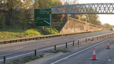 The A14 pictured with traffic cones on the road with a pedestrian bridge. 