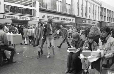 Alan Van Wijgerden A woman and two boys are seated eating food out of boxes in Birmingham city centre. Behind them a busy street with people walking outside shops including Freeman Hardy Willis, Paige Shops and Marks and Spencer 