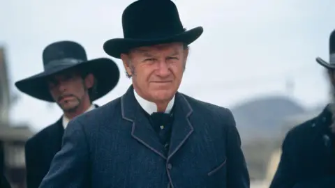 Getty Images Gene Hackman in costume - an old-fashioned blue suit and bowler hat, sporting grey sideburns - on the set of Sam Raimi's 1995 movie "The Quick and the Dead". Two other actors are seen in similar dress in the background