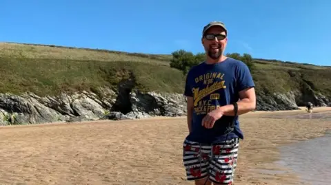The photo shows Gavin stood on a beach with a grassy cliff in the background. He smiles at the camera while wearing a blue t-shirt with yellow writing and white and blue stripped shorts. He also wears sunglasses and a baseball style cap.