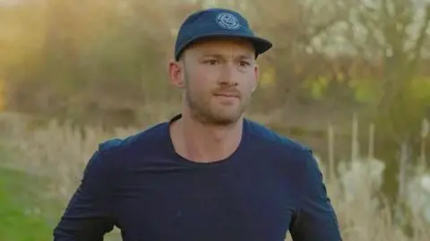 Fergus Crawley is wearing a blue top and blue cap with a closely trimmed beard and moustache. He is running in a park 