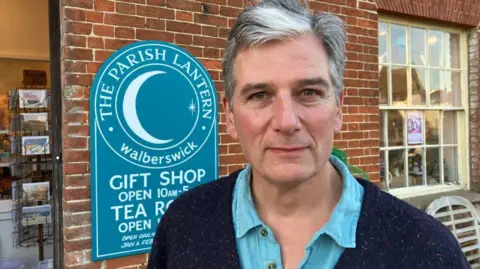 Vikki Irwin/BBC Robin McLean stands in front of his blue shop sign for The Parish Lantern gift shop and tearoom. He has grey hair, a dark blue cardigan and a light blue open-necked shirt.