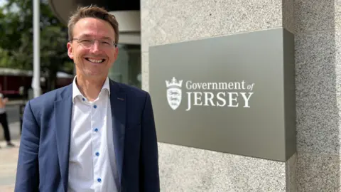 Deputy Kirsten Morel stands next to a Government of Jersey sign. He is wearing a navy blue suit jacket and a white shirt and he is smiling widely.