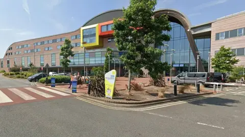 A street view of Alder Hey Children's Hospital. It is a large grey building with windows. There is a space for cars to pull into outside the entrance, and trees are dispersed across the front of the site.