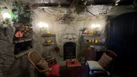 A cave like room with old-style chairs and candles, with artificial greenery.