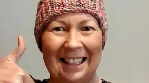 Family A woman wearing a hat is smiling at the camera in front of a plain background.