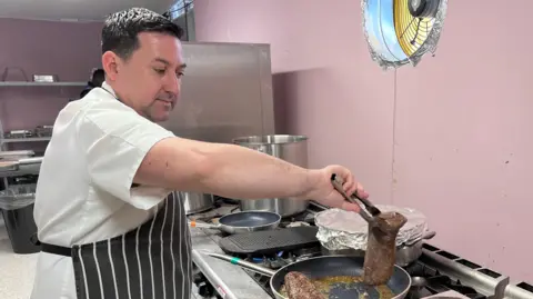 A person wearing a striped apron cooks meat in a frying pan on a commercial gas hob, using tongs to lift a piece while another cut of meat rests on a metal tray beside the stove.