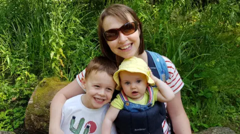 Family photo Nicola Wheatley is sitting on a  rock in a green space with her children on her lap. She and Oscar are smiling at the camera and baby Ffion is wearing a yellow summer hat and dungarees.