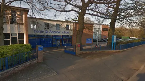 Google Thorpe St Andrew School in blue lettering above the entrance of a two-storey building. There are trees, blue metal fencing and a footpath and vehicle entrance in front of the entrance doors.