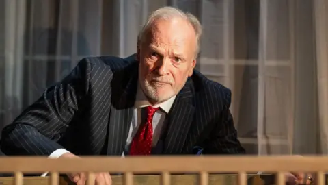 Hugo Glendinning Actor Clive Mantle looks over a bannister with a surprised expression on his face during a production of Ghost Stories at the Theatre Royal in Bath. He is wearing a dark grey pin striped suit and a red tie and white shirt