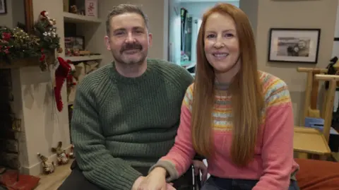 Dan and Anna smile in the living room. Dan (left) is wearing a green jumper and Anna (right) is wearing a pink winter jumper with yellow and blue patterns.
