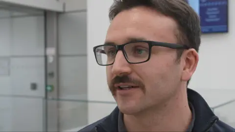 Jack Lamb is wearing black-rimmed glasses and has short, dark hair and a moustache.