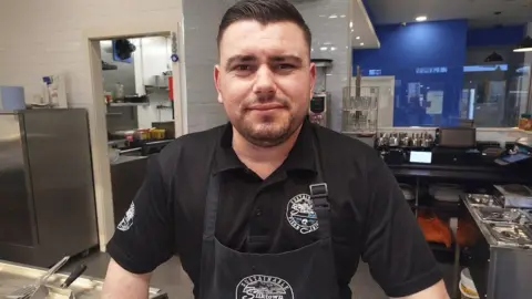 Paul Burnell/BBC Eric Meta has dark hair, beard and is wearing a black apron and shirt with his chip shop logo on them.