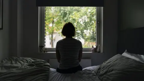Woman with short hair sat on bed looking out of window in the dark.