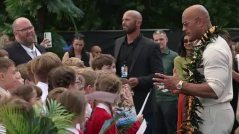 Joshua Falcon/BBC A group of schoolchildren wearing red uniforms standing in front of The Rock – a man wearing a cream jumper and white trousers and sunglasses.
