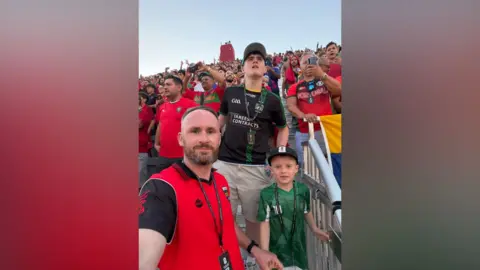 Two men and a teenager stand with the world cup tickets. The man in the red football shirt has a little boy on his shoulders. 
