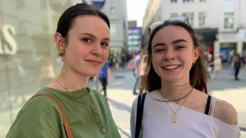 BBC Two young women look at the camera. The one on the left has brown hair tied back, wearing dangly earrings and a green coloured top. The one on the left is smiling. She has long brown hair caught back with sunglasses and is wearing a pale coloured top that is off one shoulder.
