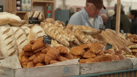 The image shows a market stall piled high with freshly baked bread, including loaves, baguettes and croissants, arranged in wooden trays with small price signs such as “Plain Croissant £3”. A person wearing a cap stands behind the counter serving customers, while a crowd of shoppers fills the background.