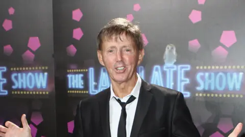 Getty Images Dickie Rock with short brown hair and wearing a black suit and white shirt with a neon Late Late Show sign in the background