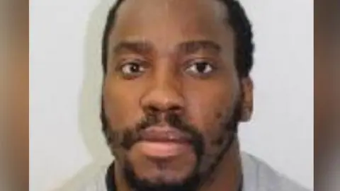 Met Police Kamar Williams. He has close cropped black hair and a beard. He is wearing a grey top
