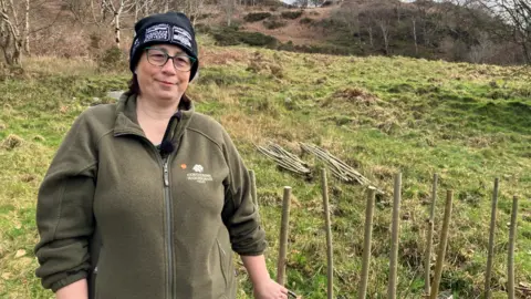 A woman is standing on a hillside. She's wearing spectacles and a black beaning. She's also wearing a khaki green fleece which has the Woodland Trust logo on it.