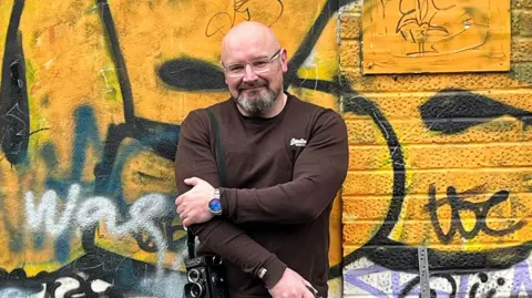 Colin Howey Stuart McPherson standing in front of a graffiti-painted wall. He has his arms crossed and is looking directly at the camera and smiling. He is wearing a brown long-sleeved T-shirt.