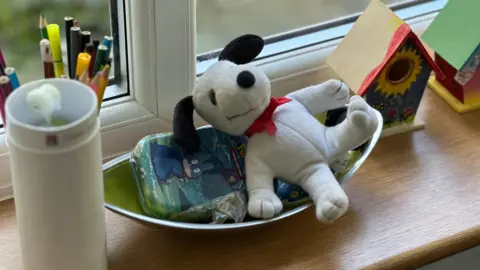 BBC Close up of a wooden window ledge, taken in a children's home. On the ledge are a plastic container, some felt pens and coloured pencils, a bowl containing a pencil case and a cuddly toy, and two painted wooden toys.
