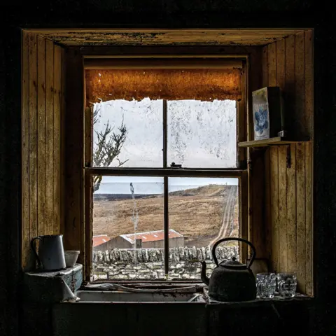 Angus Mackay Photography Looking out through a window of the abandoned house. The window has a tattered blind. There is a sink and on it an old kettle.