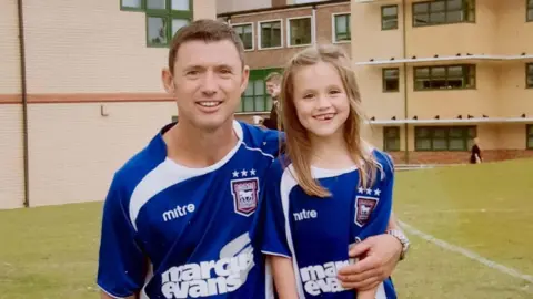 Supplied Chris is wearing an Ipswich Town shirt, on one knee, with his arm around a young Eloise King, who is draped in an Ipswich Town shirt that is charmingly, slightly too big. They are both smiling. 