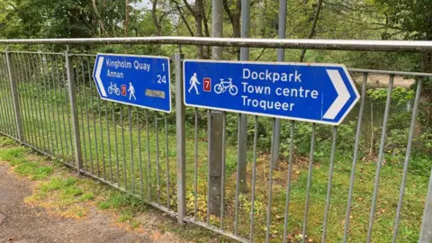 Signs pointing in opposite directions. One points to Dockpark, the town centre and Troqueer. The other to Kingholm Quay and Annan. 