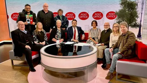 Tom Hedges in a dark suit sits on the left of a curved red sofa in the BBC Breakfast TV studio. Around him are other wrongly accused sub-postmasters and the programme's two presenters.