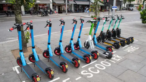 Dominic Lipinski/PA Wire A row of e-scooters, all facing away from the camera, parked in a bay marked out with white lines in a town centre.  The writing on the pavement around the parking bay reads "e-scooter hire"