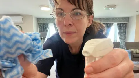 Catriona MacPhee working as a cleaner at the care home. She is wearing glasses, her hair is tied back and she is holding a spray bottle and cloth. 