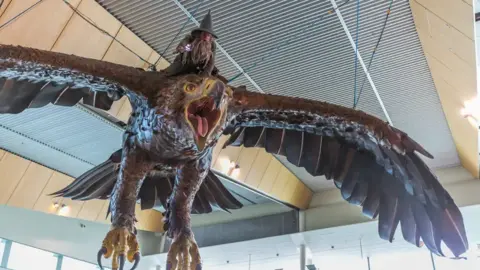 Wellington Airport A shot from the ground of a giant eagle sculpture hanging from the ceiling, with its wings spread out and its mouth open. Gandalf is perched on the back of the sculpture.