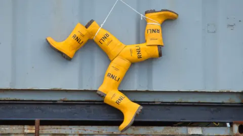 An Isle of Man triskelion made of several pairs of yellow RNLI wellies is hanging from the wall of a shipping container