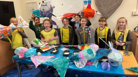 A group of young female pupils stand in a line, wearing brown and yellow brownies uniform. In front of them lies a table covered in colourful fish-shaped lanterns, along with black trays filled with PVA glue and spreaders.