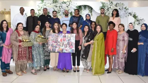 Love in Care Ltd Image shows a group of people celebrating 10 years since Love in Care's launch. Aqila Choudhry is featured in the centre and is wearing a purple dress and is holding a photo frame. Other employees are gathered around her.
