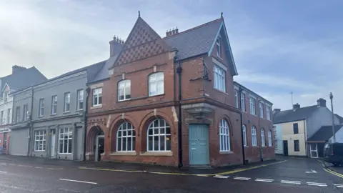 A red bricked building sits at the corner of the road. It has a green double door, and semi circle shaped windows on the ground floor. It looks like a historical building. 