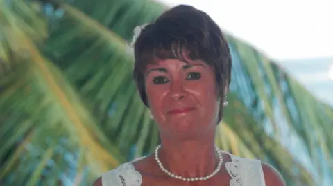 A woman smiling with short brown hair, wearing a white dress. In the background are palm trees.