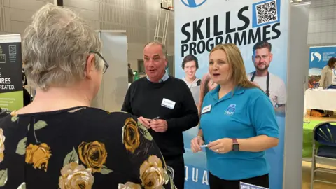 Shows a woman in a dark floral dress, a man in a dark jumper and a woman in a blue t-shirt in front of a banner saying "skills programmes".