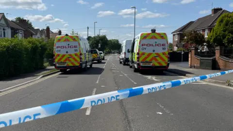 A police cordon tape across a residential street with police vans in the background