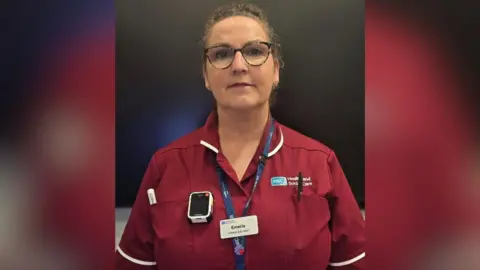BBC A woman with curly brown hair tied back from her face. She is photographed from the waist up, against a black background, wearing black glasses and a red HSC nurses top. A white body camera is visible on her uniform, as is a lanyard with her name, Emeria.
