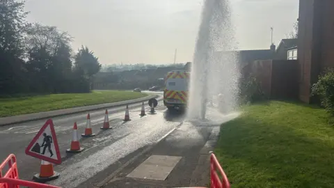 Water is seen shooting into the sky with a Severn Trent van parked nearby and cones marking out the affected area.
