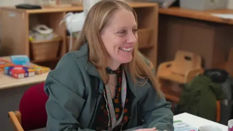 Ant Saddington/BBC Diane sits at a table in a community centre, looking at someone on the other side (who we cannot see), smiling at them with a cheerful smile. She wears a colourful lanyard around her neck and an open green jacket over a black blouse.
