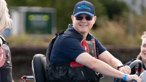 Nathanial Rosa Paul Wilkinson on a motored water vehicle, wearing a floatation vest and sunglasses. He is smiling.