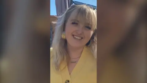 A woman with blonde hair and a yellow cardigan smiles directly to the camera 