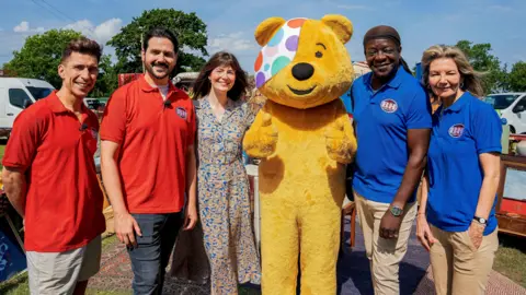 A group shot of Pudsey Bear standing in the sunshine in a car park in a field surrounded by five people, one pair in red polo shirts and one pair in blue polo shirts. Natasha Ruskin Sharp, the Bargain Hunt presenter has her arms around Pudsey