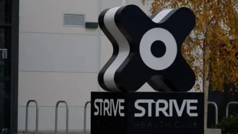 The outside of the Strive health and fitness centre in St Peter, Jersey. The large 'x' shaped branded Strive sign is visible in the foreground. A grey building is visible in the background.